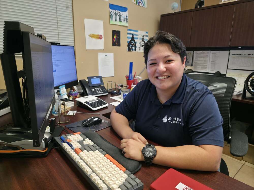 A woman wearing a Winter Park Housing branded shirt sitting at a desk in an office smiling for a photo.