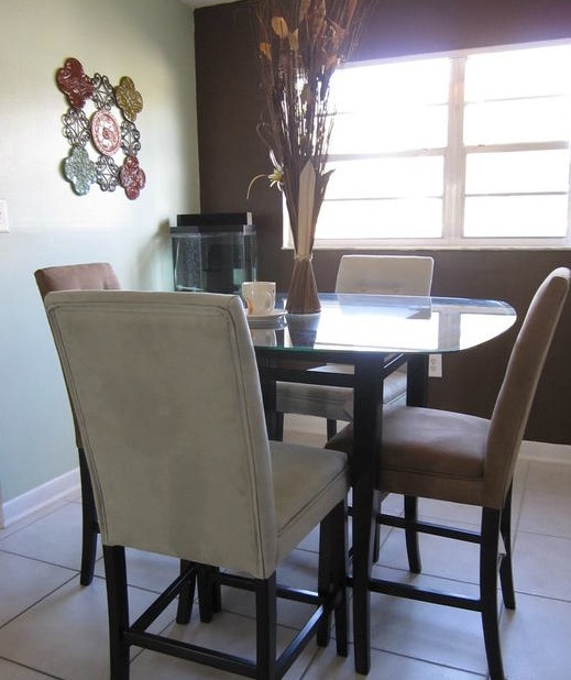 Sun streams in the window reflecting off the dinner table that has 4 chairs pulled up to it. 