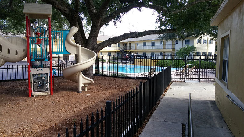The playground equipment is enclosed by a black fence and sits next to the pool, which is also enclosed by a black fence. 