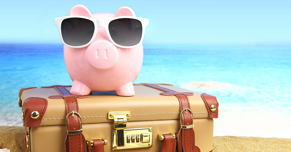 A piggy bank with sunglasses on laying on a suitcase on a beautiful beach.