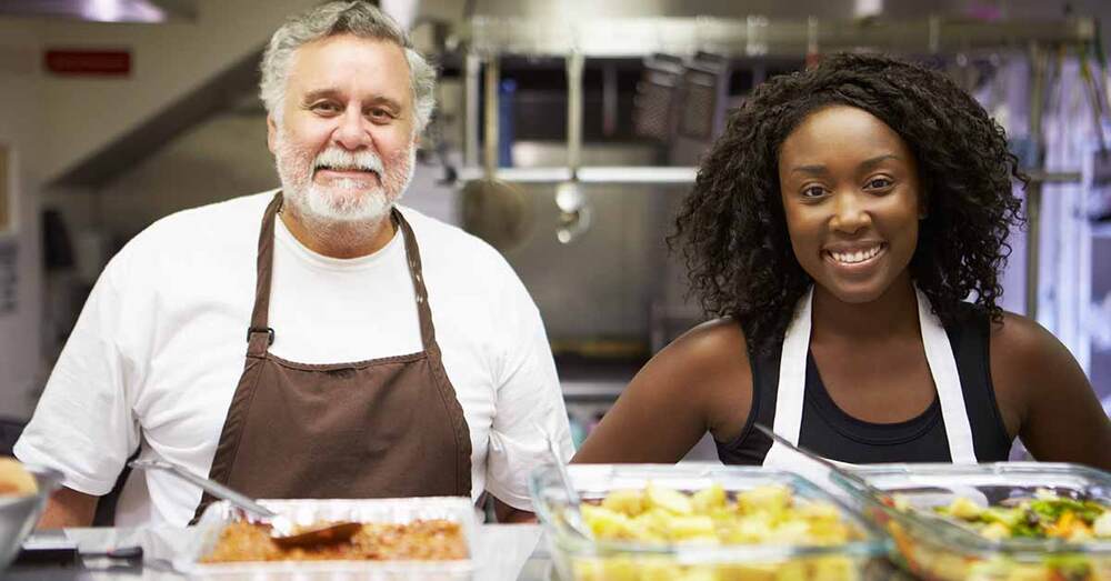 Two individuals standing in a kitchen in front of freshly cooked food.