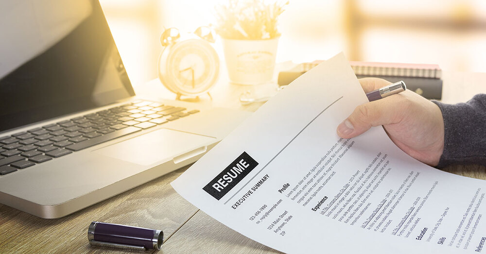 An individual holding a resume by a laptop.