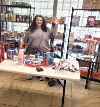 lady behind a table and in front of shelves covered with items in the food pantry.