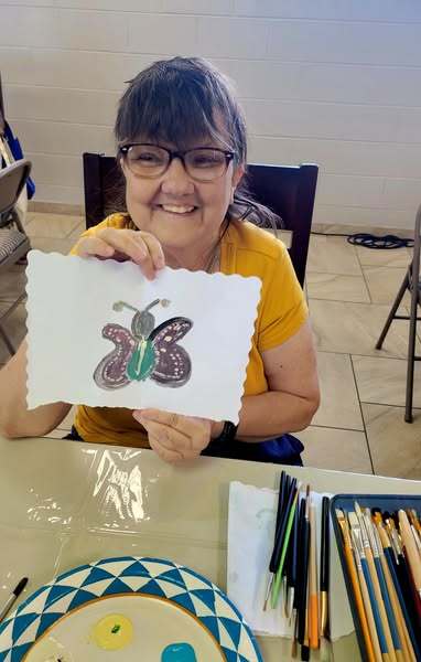 A woman smiling and holding up a picture of a butterfly she drew.