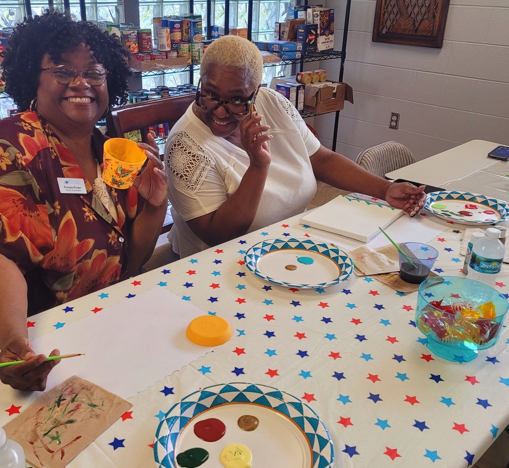 Two women sitting at a table, smiling while making art.