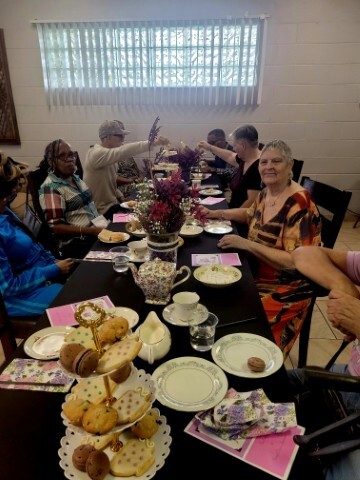 Men and women smiling and talking around a table with cookies and teacups with saucers. 