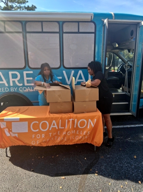 A couple ladies unpacking boxes of supplies on a table in front of a van.
