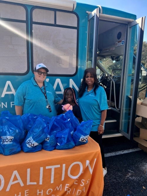 People in a row behind a table full of supplies in front of a van.
